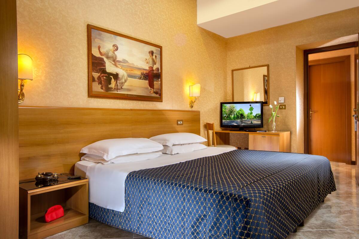 A hotel room with a large bed, a television, a painting on the wall, and a wooden headboard.