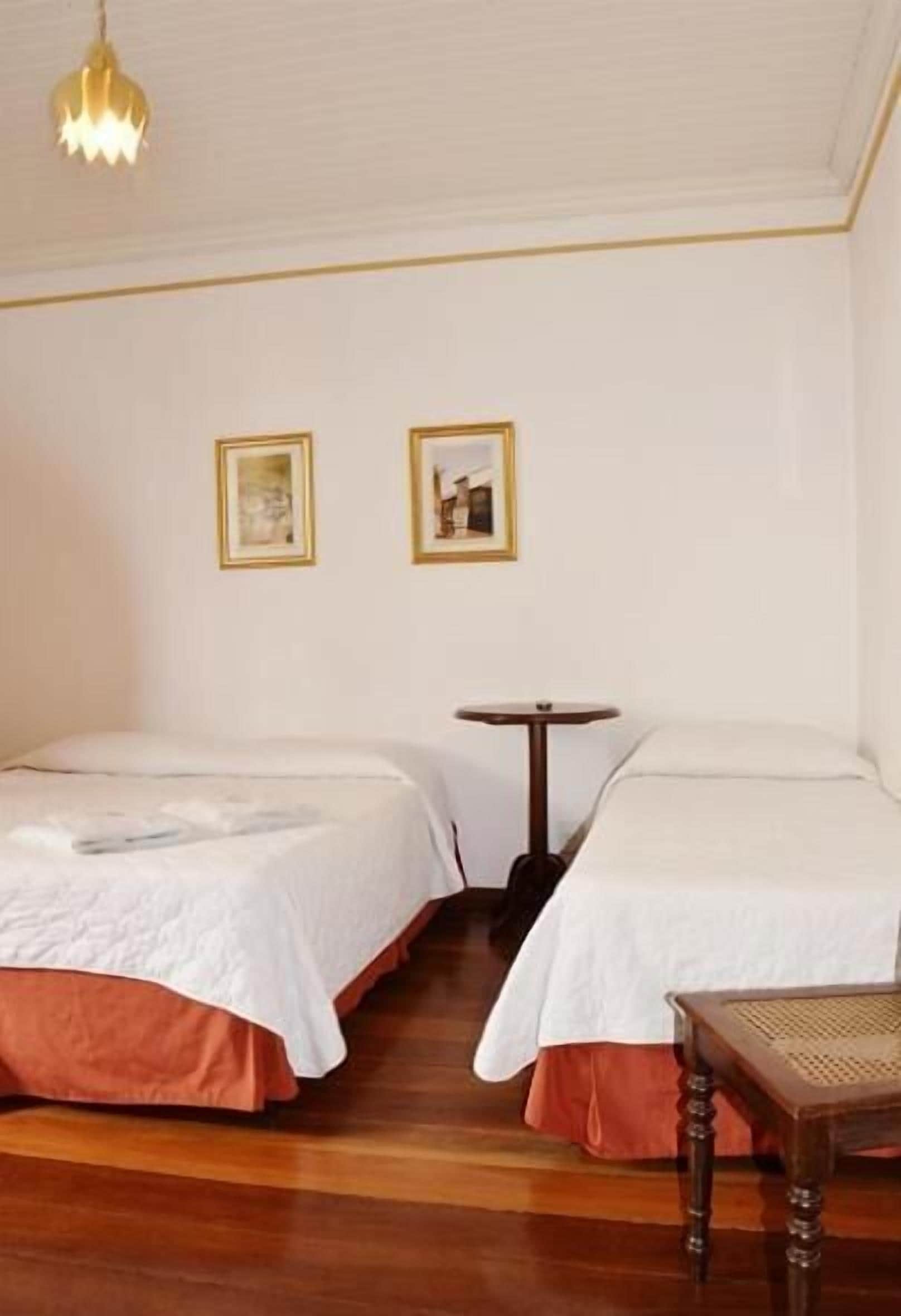 Ein Zimmer mit zwei Betten, einem kleinen runden Tisch und zwei gerahmten Bildern an der Wand.