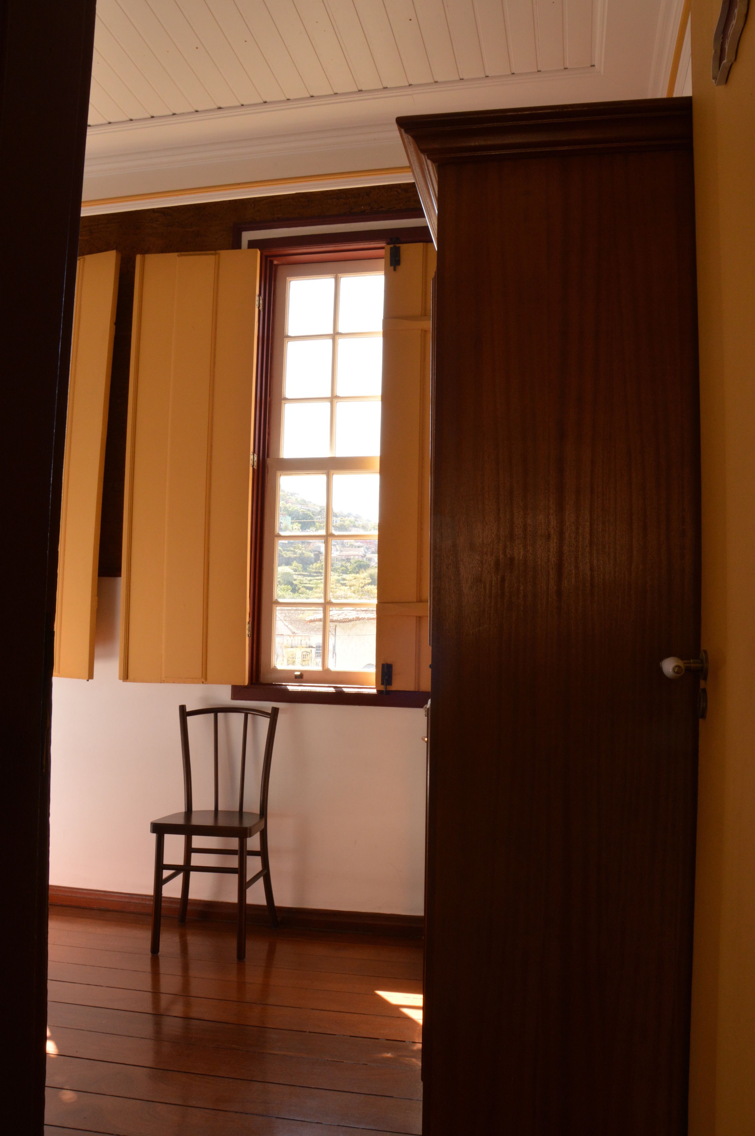 Una habitación con suelo de madera, una ventana con contraventanas amarillas y una puerta de madera.