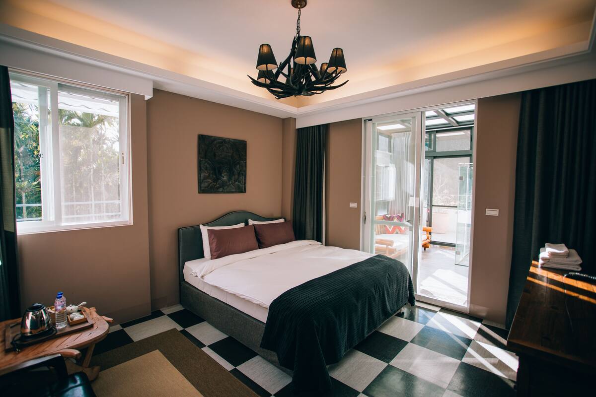A hotel room with a checkered floor, a bed with a dark headboard, a chandelier, and a view through a glass door.