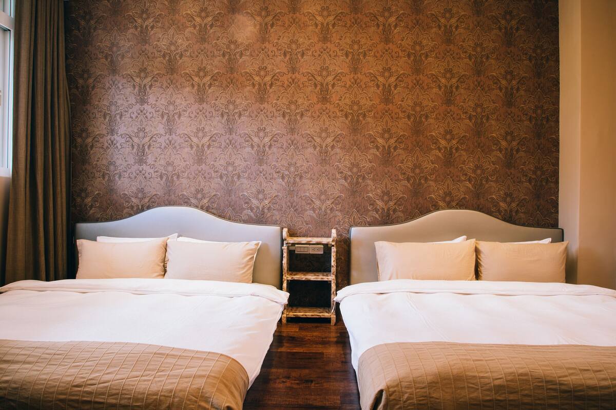 A hotel room with two beds, a wooden nightstand, and a patterned wallpaper.
