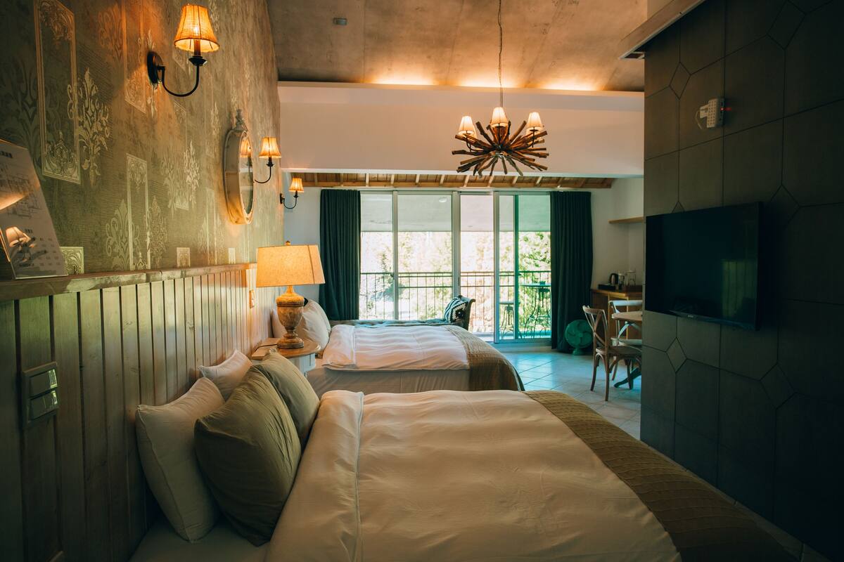 A hotel room with two beds, a TV, a dining area, and a view of the outdoors.