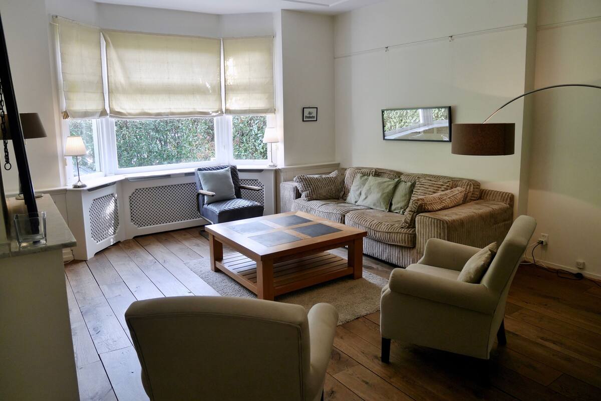 Ein Wohnzimmer mit Holzboden, einer Couch, einem Sessel, einem Couchtisch und einem Fenster mit Vorhängen.