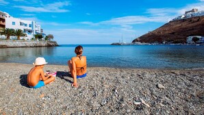 Una spiaggia di ciottoli con due persone sedute, che guarda un mare calmo e una cittadina costiera con edifici bianchi.