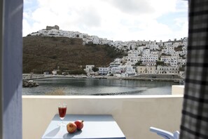 A balcony with a table, a glass of red drink, and two pomegranates. In the background, there is a hillside with white buildings and a body of water. - Vithos Aparthotel (Astypalaia)