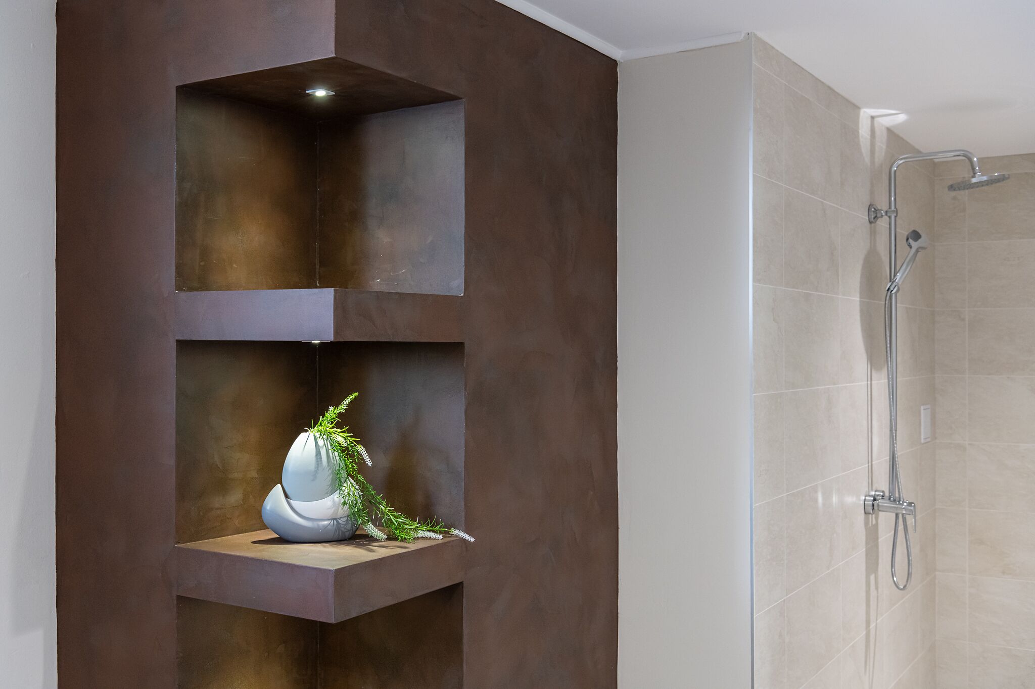 A modern bathroom with a built-in shelf holding a decorative vase and greenery, and a separate shower area with a handheld showerhead.