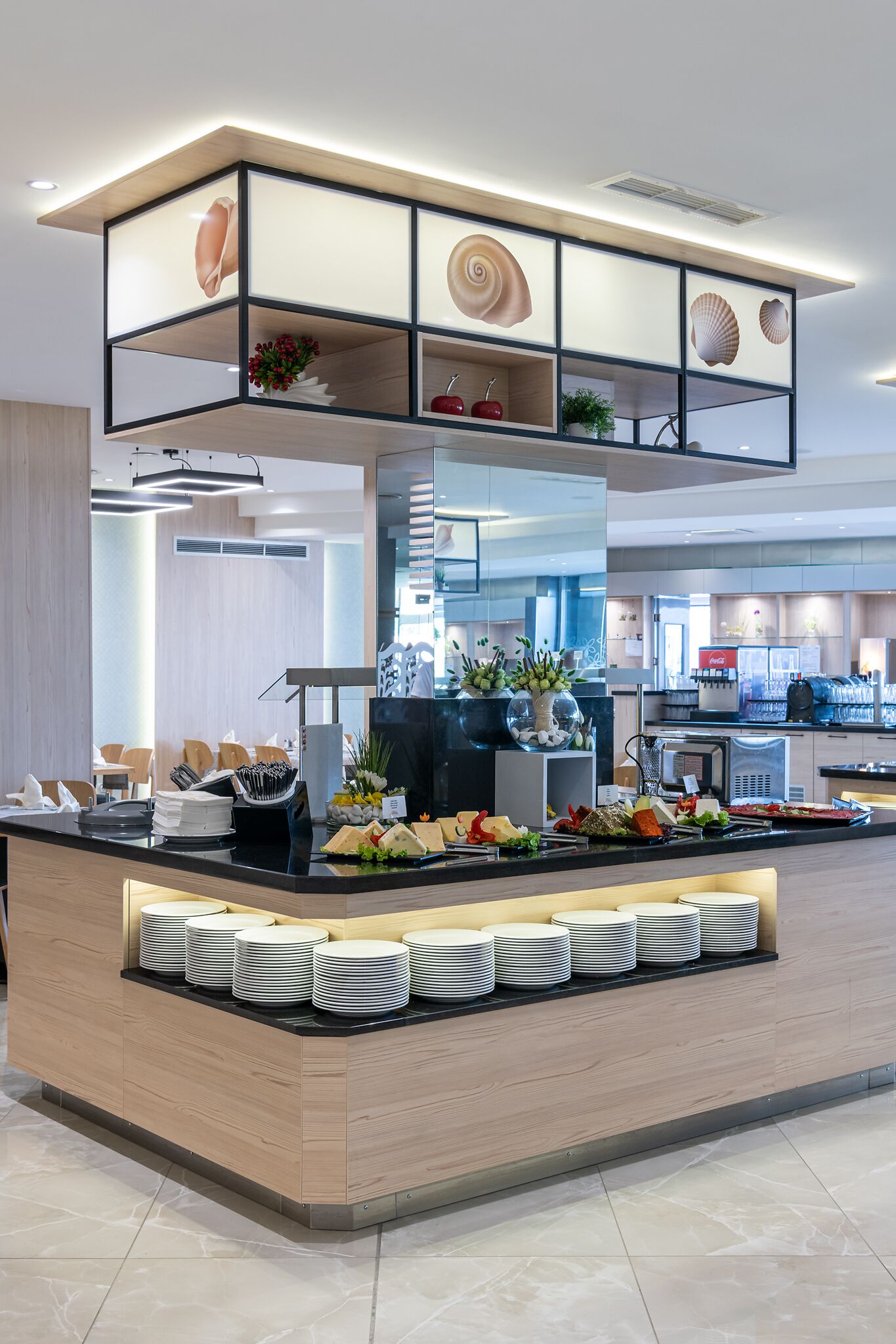 A modern hotel lobby with a central reception area featuring a wooden counter, a glass display case with decorative shells, and a variety of food and drink options.