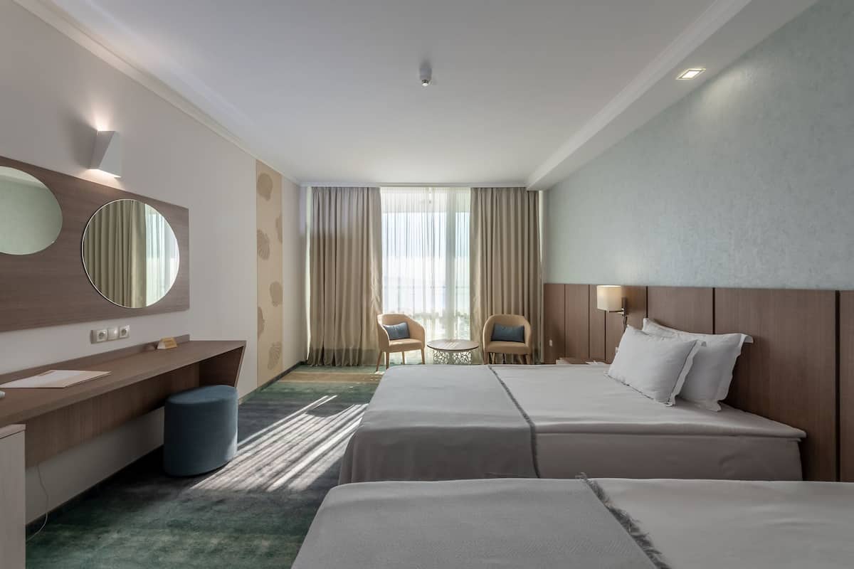A hotel room with two beds, a desk, a chair, a round table, and a large window with curtains.