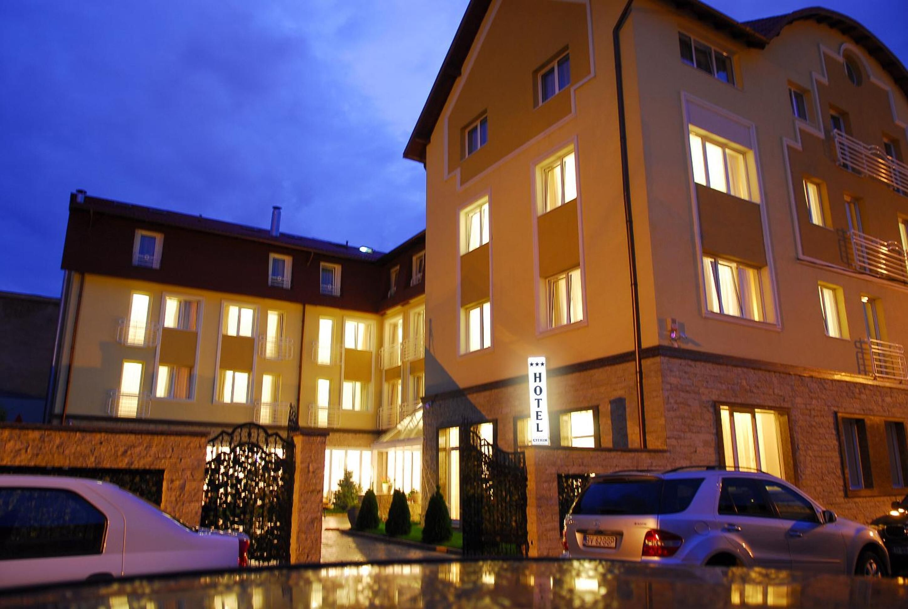 A hotel with multiple floors, a well-lit entrance, and a parking area with several cars.