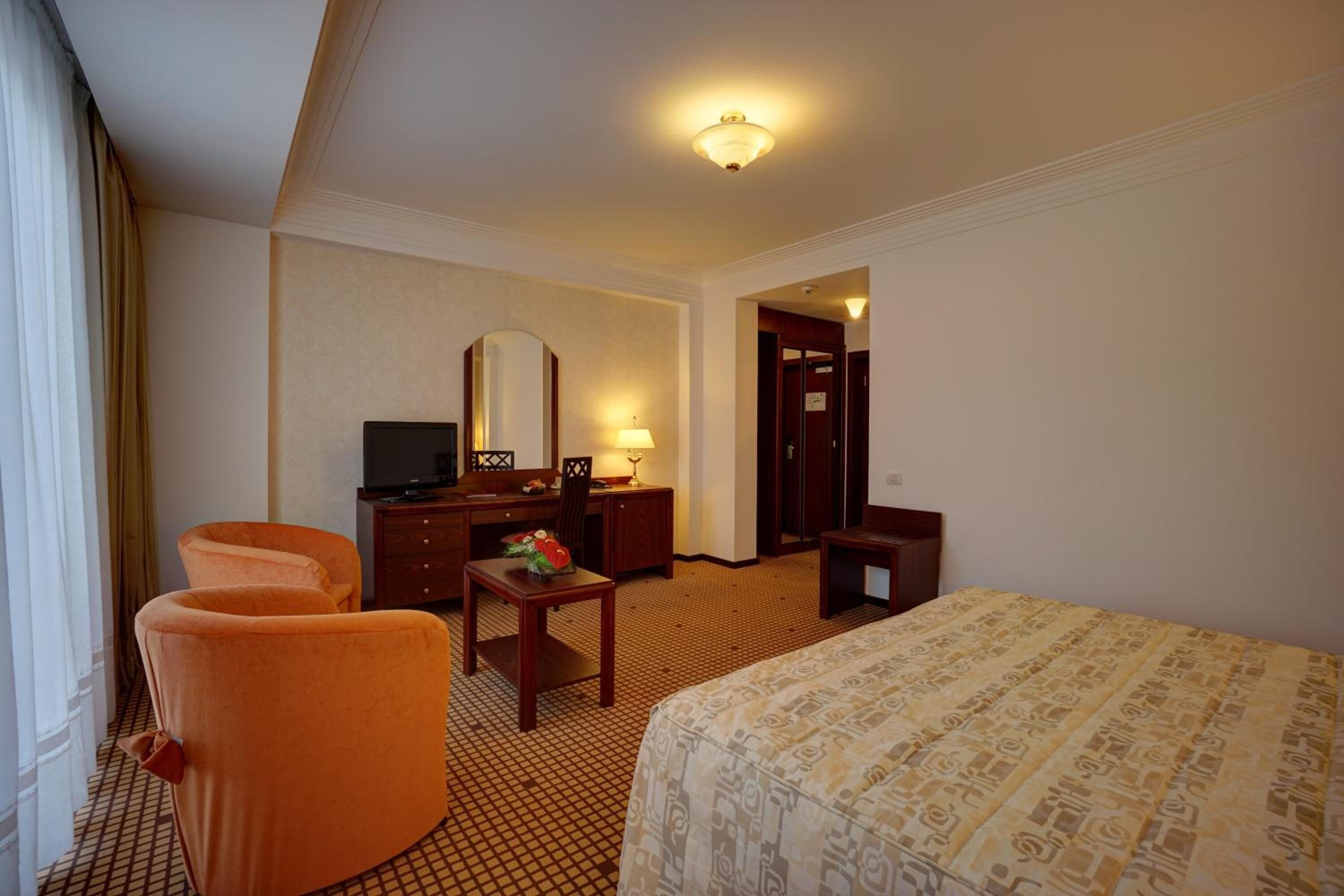 Habitación de hotel con una cama, dos sillones naranjas, un escritorio con espejo, un televisor y una pequeña mesa con una flor.