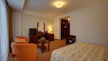 Habitación de hotel con una cama, dos sillones naranjas, un escritorio con espejo, un televisor y una pequeña mesa con una flor.