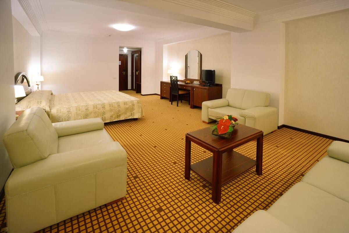 A hotel room with a bed, two sofas, a desk with a chair, a television, and a small table with a flower vase.