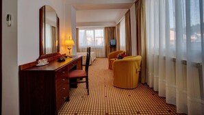 Una habitación de hotel con piso a cuadros, un escritorio de madera con una lámpara, una silla, una cama con cubrecama blanco, un televisor y ventanas grandes con cortinas.