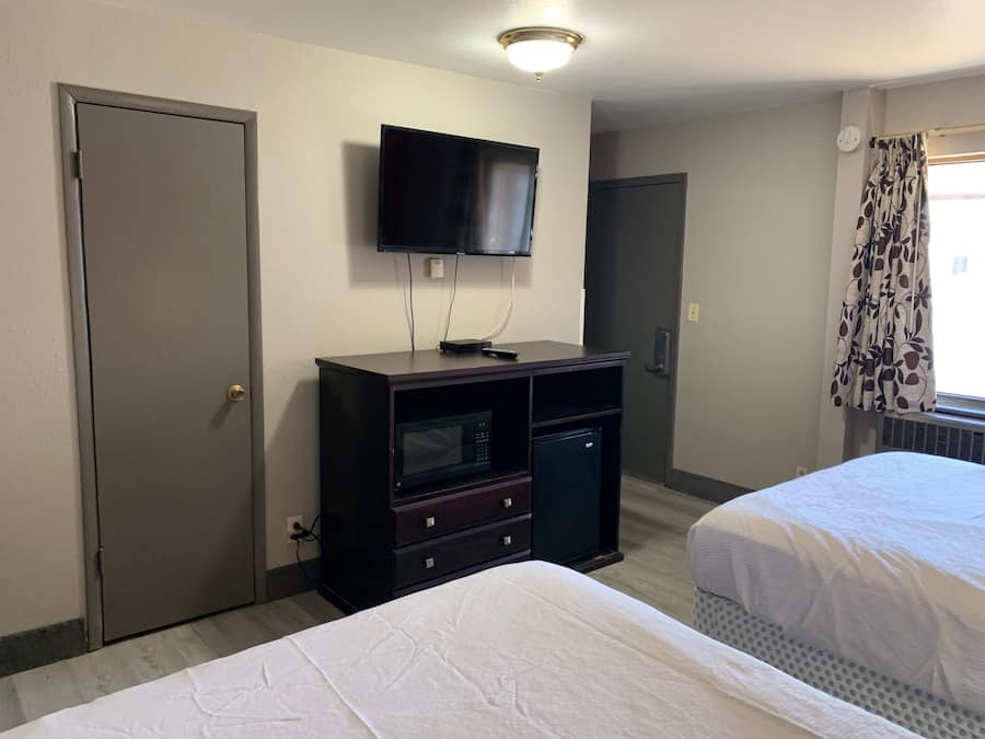 Quarto de hotel com duas camas, uma TV fixada na parede, uma secretária pequena com gavetas e uma porta.