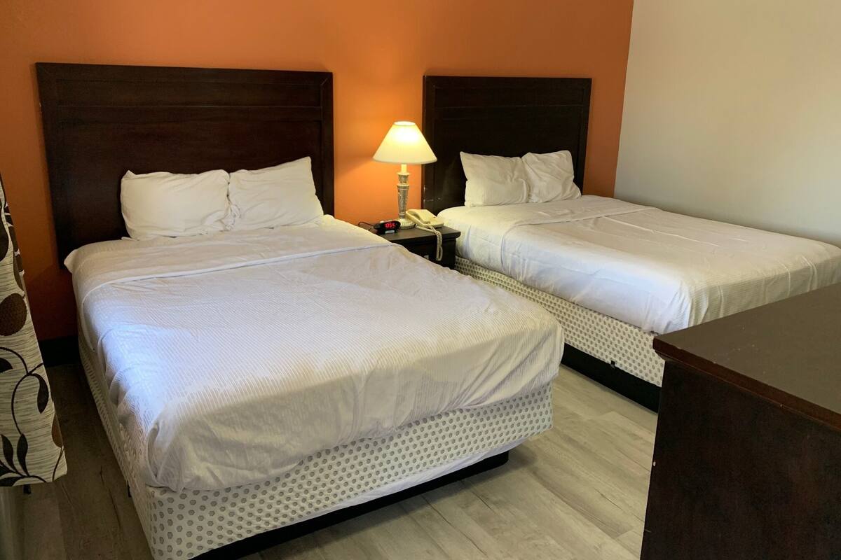 A hotel room with two beds, each with white bedding and a wooden headboard. There is a nightstand with a lamp and a telephone between the beds. The room has an orange wall and wooden flooring.