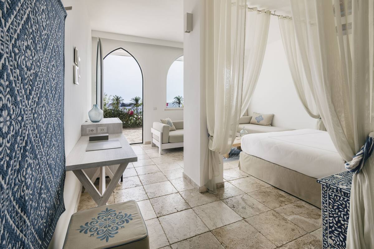 A bedroom with a large bed, a sitting area with a sofa, a small table, and a view of the outdoors.