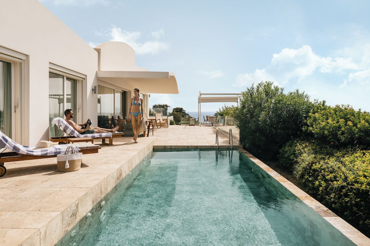 A pool area with lounge chairs and a pergola, surrounded by greenery and a clear sky.