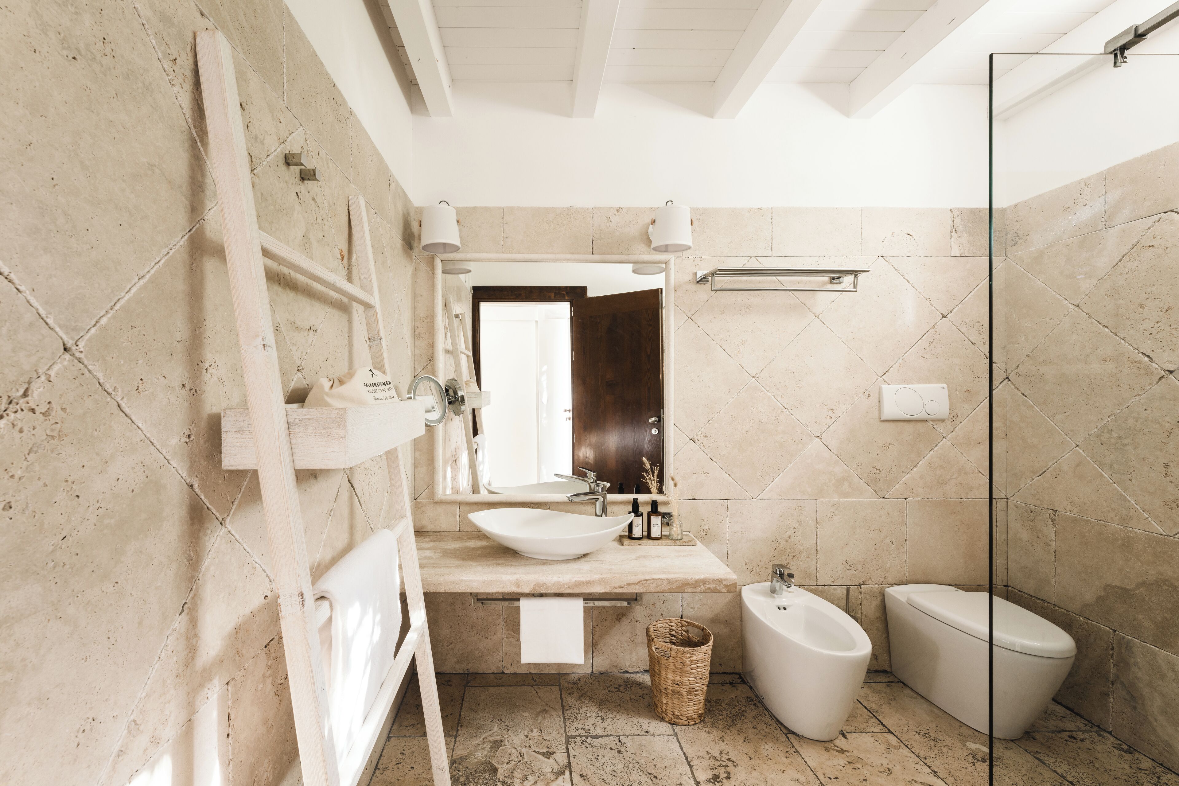 A modern bathroom with a large vessel sink, a bidet, and a toilet. There is a wooden ladder and a wicker basket on the floor.