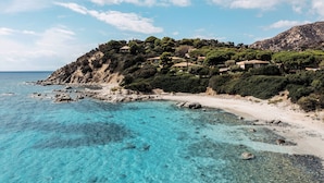 A coastal landscape with clear turquoise waters, a sandy beach, and buildings on a hillside. - Falkensteiner Resort Capo Boi (Villasimius)
