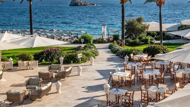 A seaside restaurant with outdoor seating, palm trees, and a view of the ocean. - Falkensteiner Resort Capo Boi (Villasimius)