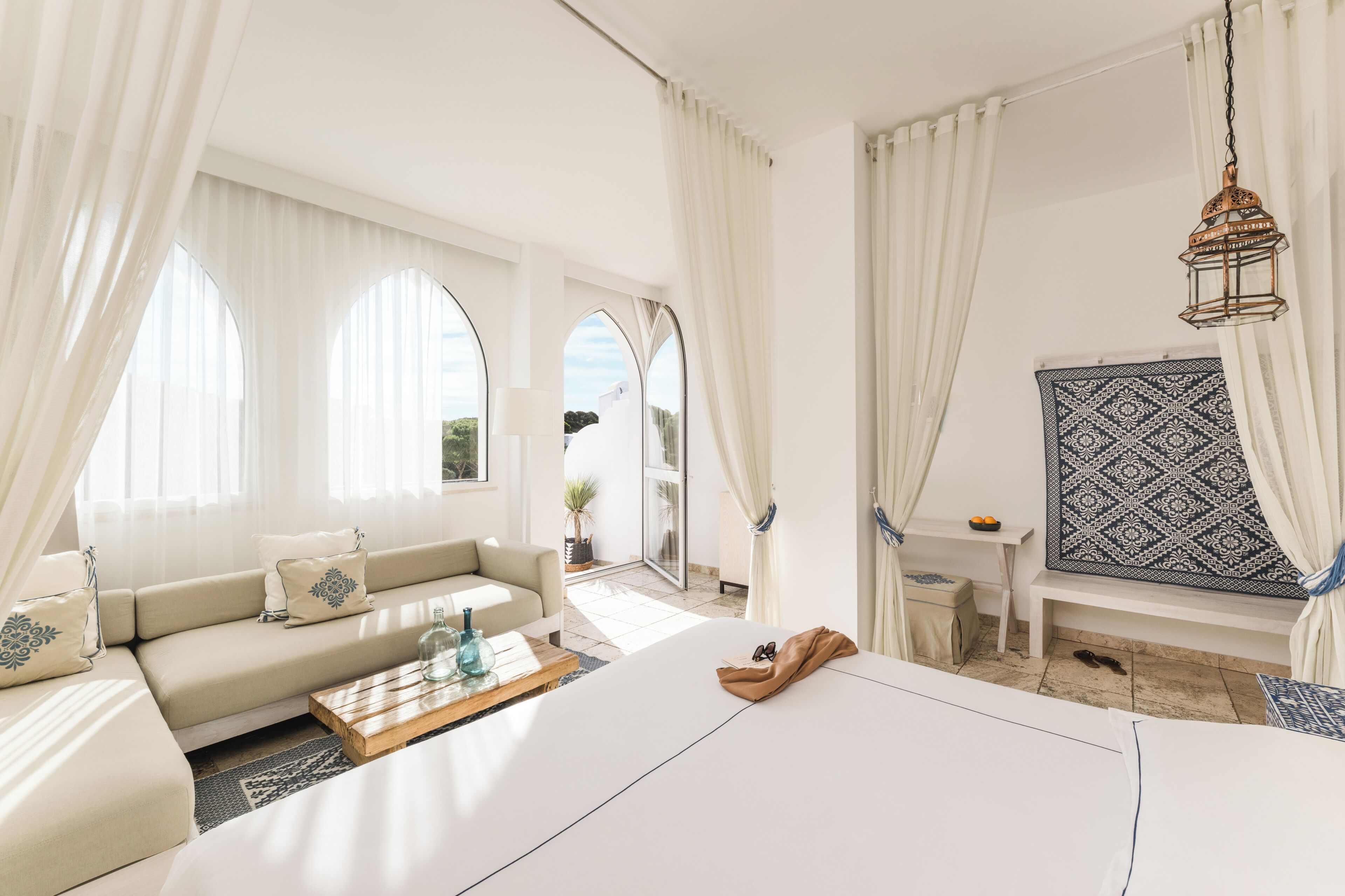 A modern hotel room with a sofa, a wooden coffee table, a bed, a small table, and a view of the outdoors.