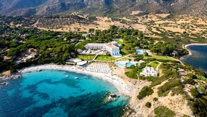 An aerial view of a coastal resort with a beach, clear turquoise waters, and mountainous backdrop. - Falkensteiner Resort Capo Boi (Villasimius)