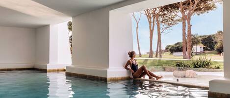 Zona de piscina con agua azul cristalina, una persona sentada junto a la piscina y vistas a árboles y césped.