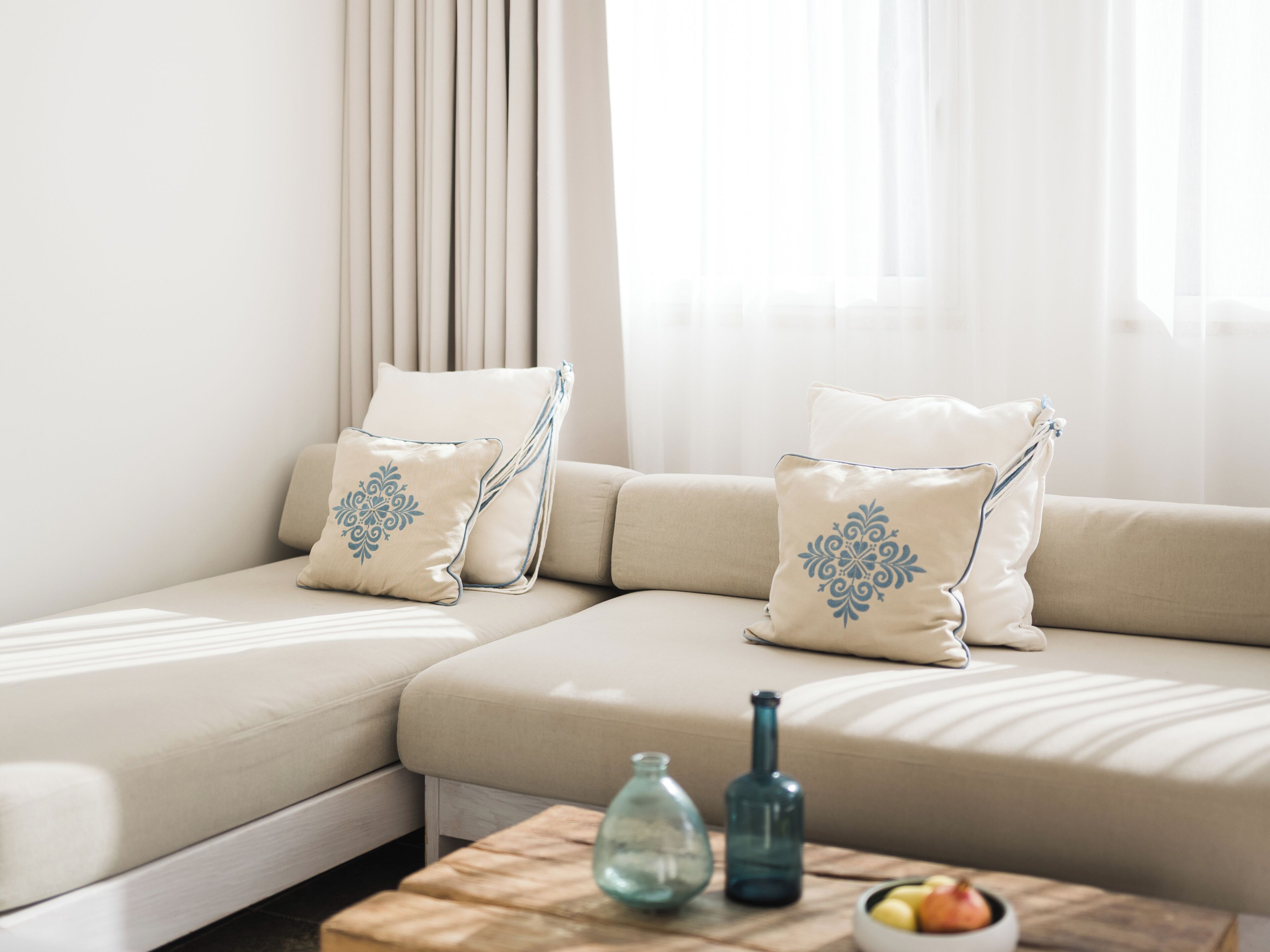 Una sala de estar moderna con un sofá seccional color beige, dos cojines decorativos, una mesita de centro de madera con una botella de vidrio y un cuenco de fruta.