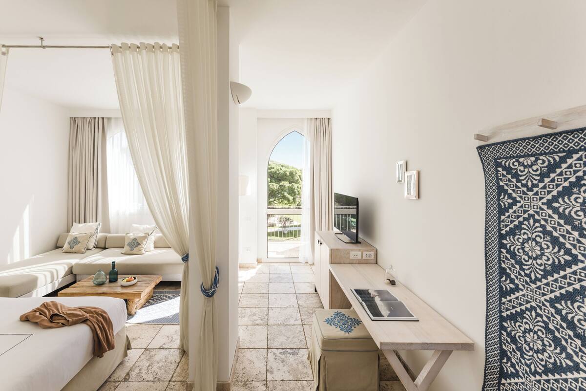 A bedroom with a bed, a desk, a TV, and a view of a garden.