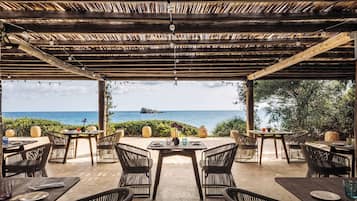 An outdoor dining area with wooden tables and chairs, overlooking the sea.