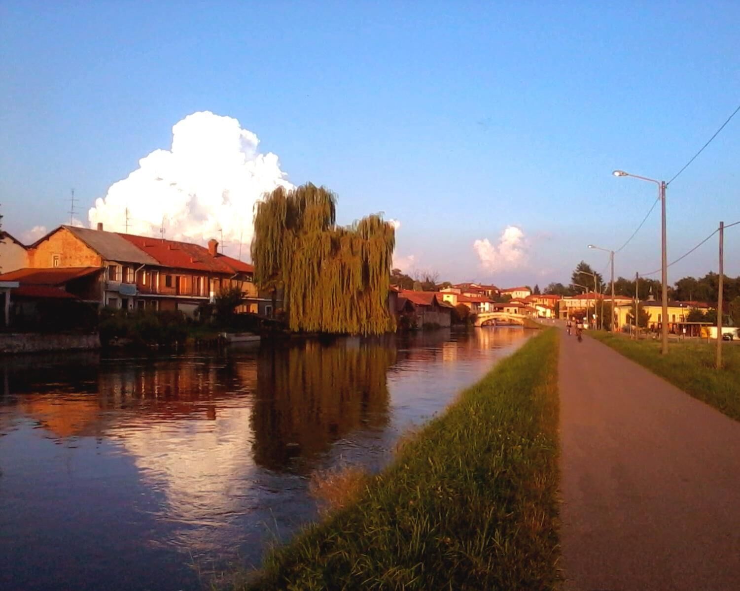 Een pad langs de rivier met gebouwen aan de ene kant en een treurwilg die in het water wordt weerspiegeld.