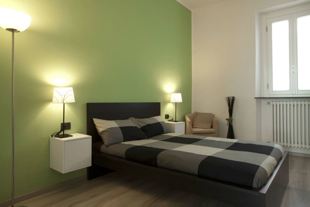 A bedroom with a bed, two bedside lamps, a nightstand, a chair, and a window with blinds.