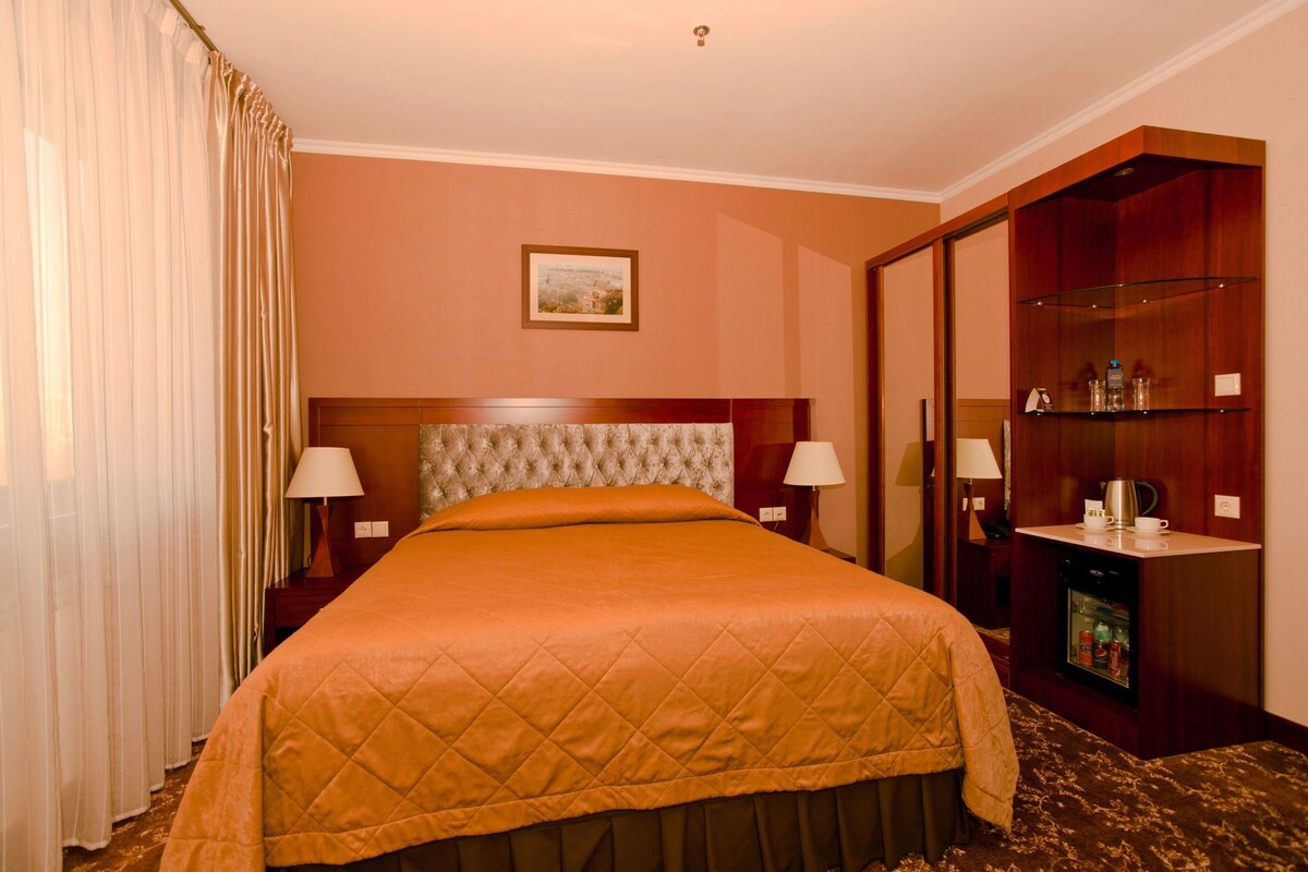 A hotel room with a large bed, two bedside lamps, a wooden headboard, a framed picture on the wall, a minibar, and a wardrobe.