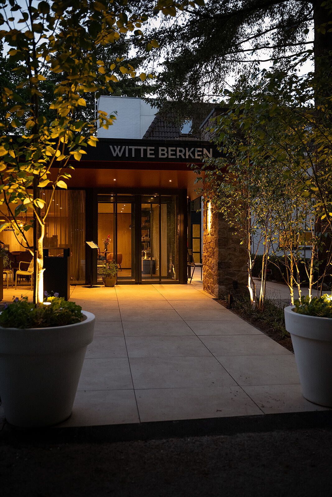 Un edificio con un letrero que dice 'WITTE BERKEN' en la entrada, rodeado de plantas en macetas y un camino pavimentado.