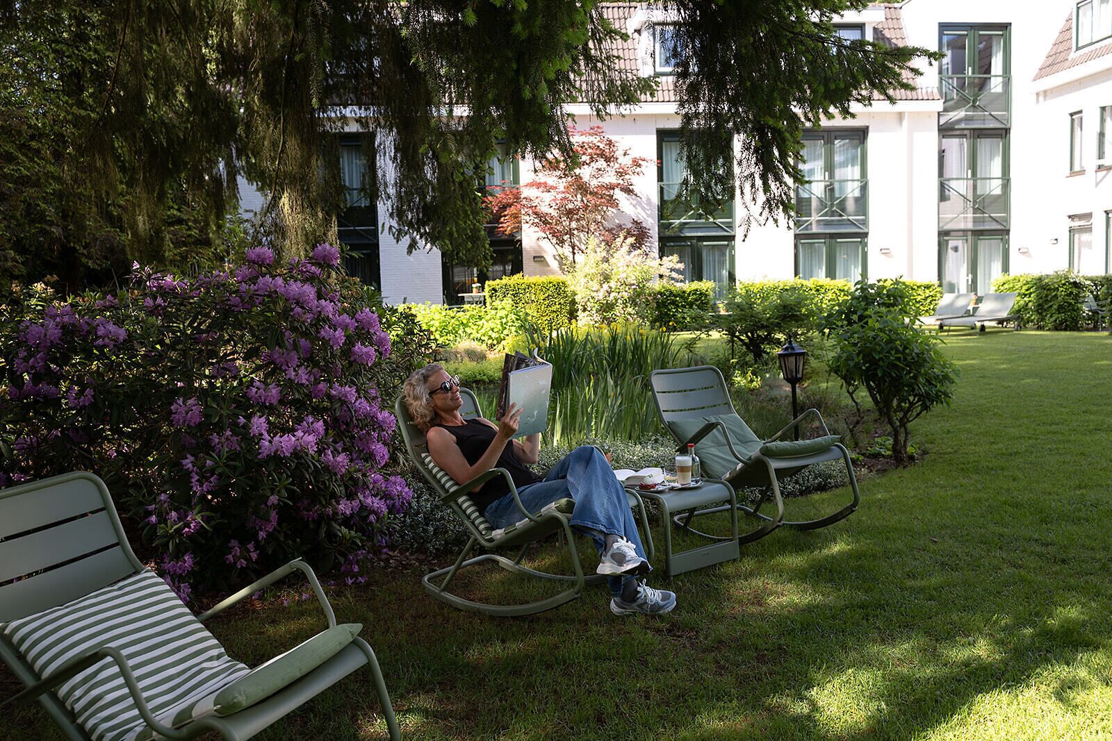 Eine Person entspannt in einem Stuhl in einem gepflegten Garten mit Blick auf ein Gebäude im Hintergrund.