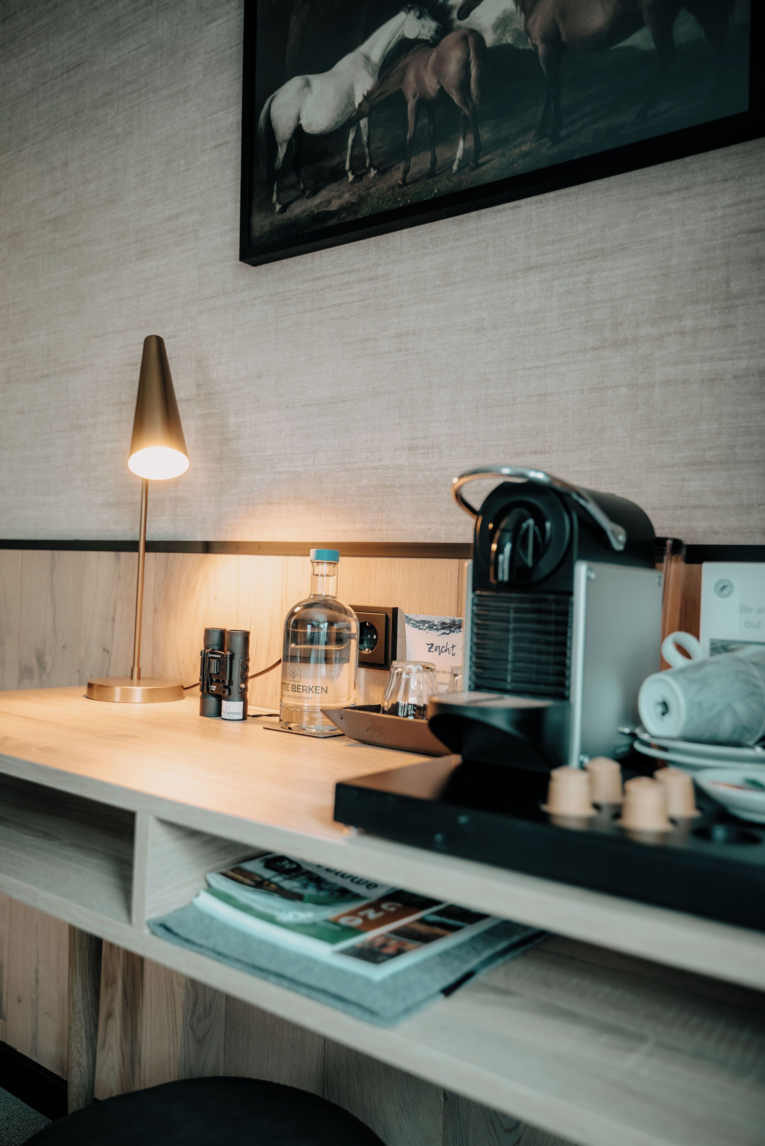 Habitación de hotel con un escritorio de madera, una lámpara, una botella de agua, una radio y un cuadro en la pared.