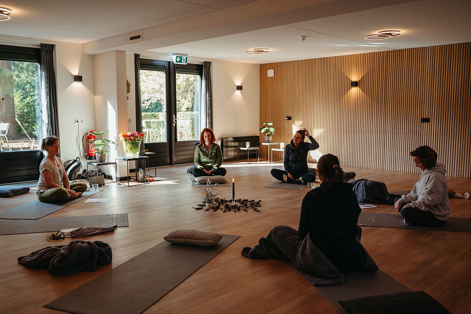 A yoga class in a room with wooden floors, large windows, and a view of greenery outside.