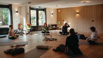 A yoga class in a room with wooden floors, large windows, and a view of greenery outside.
