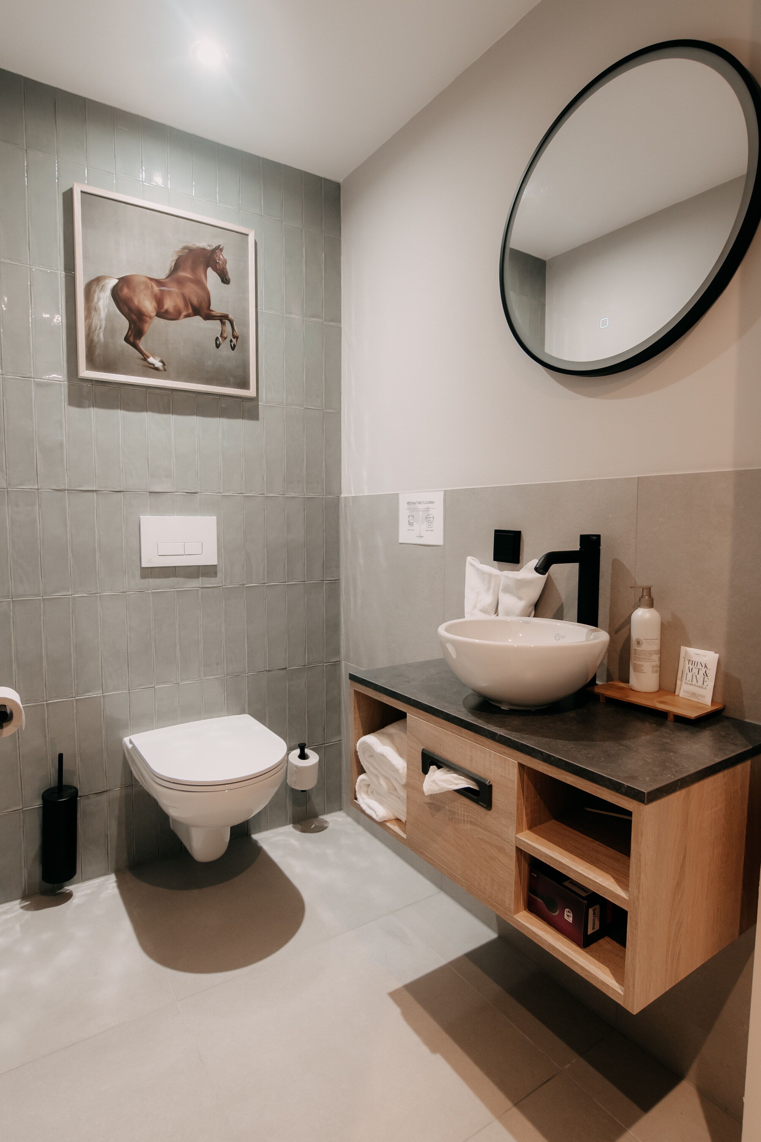 Een moderne badkamer met een grote ovale spiegel, een schilderij van een paard in een lijst, een witte wasbak en een houten wastafelmeubel met opbergruimte.