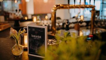 Zona de bar con un cartel que dice 'botanist drinks & bites', un florero de vidrio con una planta y un fondo desenfocado con luces.