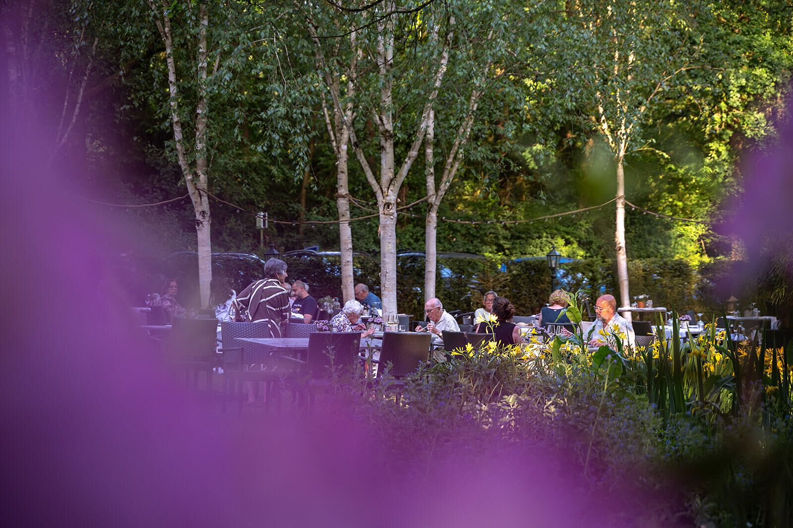 An outdoor dining area with tables and chairs, surrounded by trees and greenery.