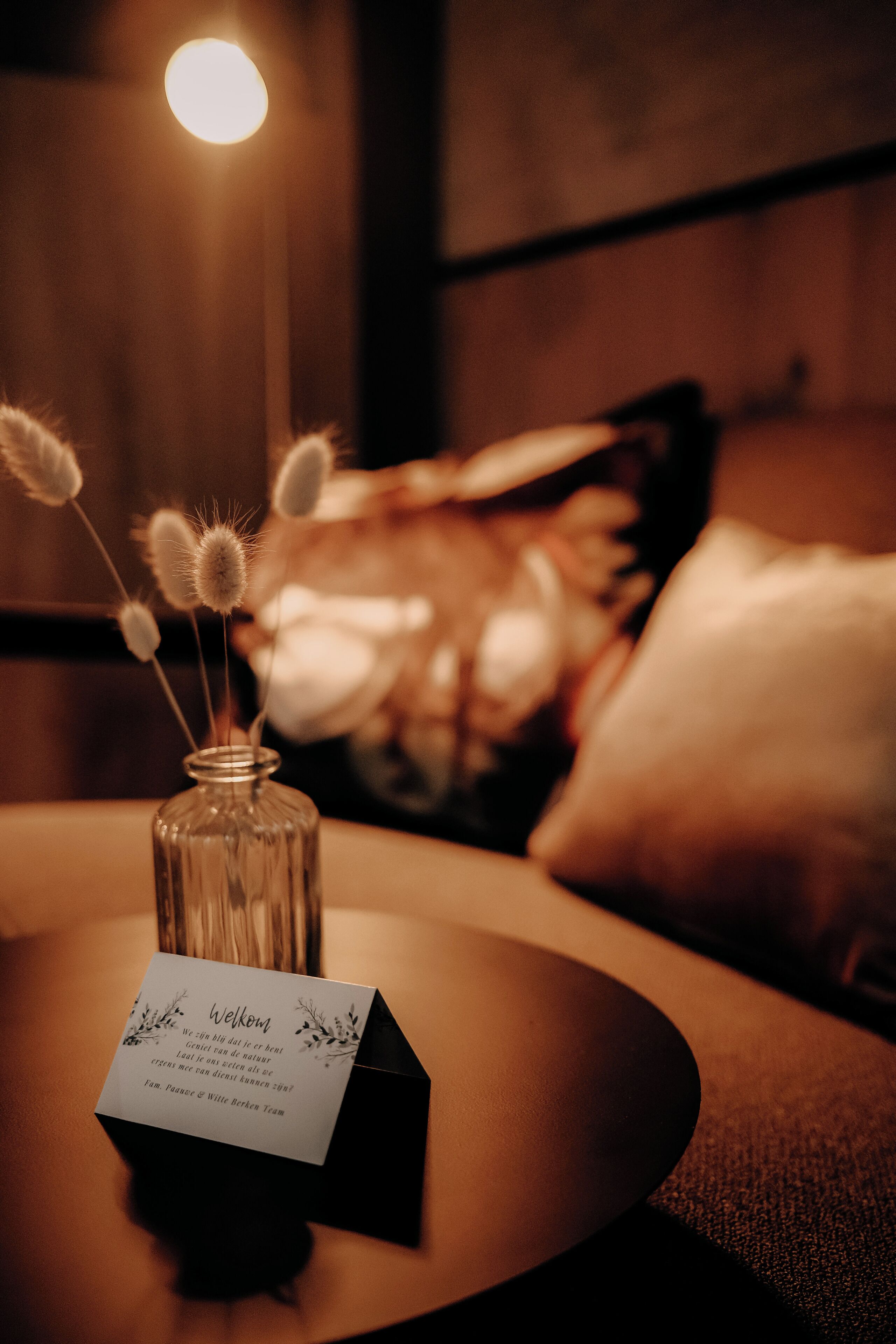 Un jarrón de vidrio con flores secas sobre una mesa, una tarjeta de nombre y una habitación tenuemente iluminada con una pared de madera y un cojín al fondo.