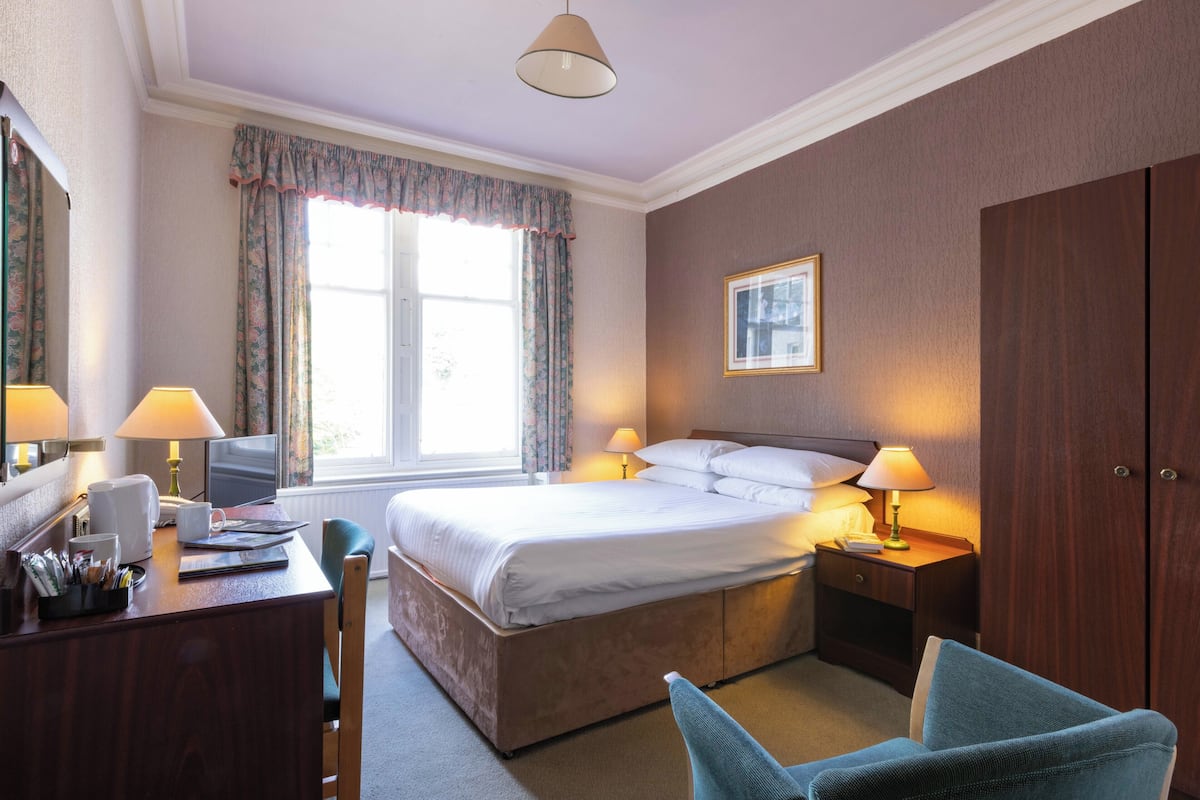 Quarto de hotel com uma cama grande, duas luminárias de cabeceira, uma escrivaninha com luminária, uma cadeira, um guarda-roupa e uma janela com cortinas.