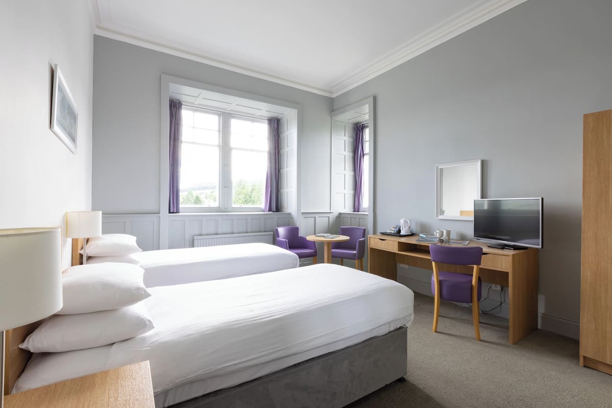 Quarto de hotel com duas camas, uma escrivaninha com computador, uma cadeira e uma televisão.