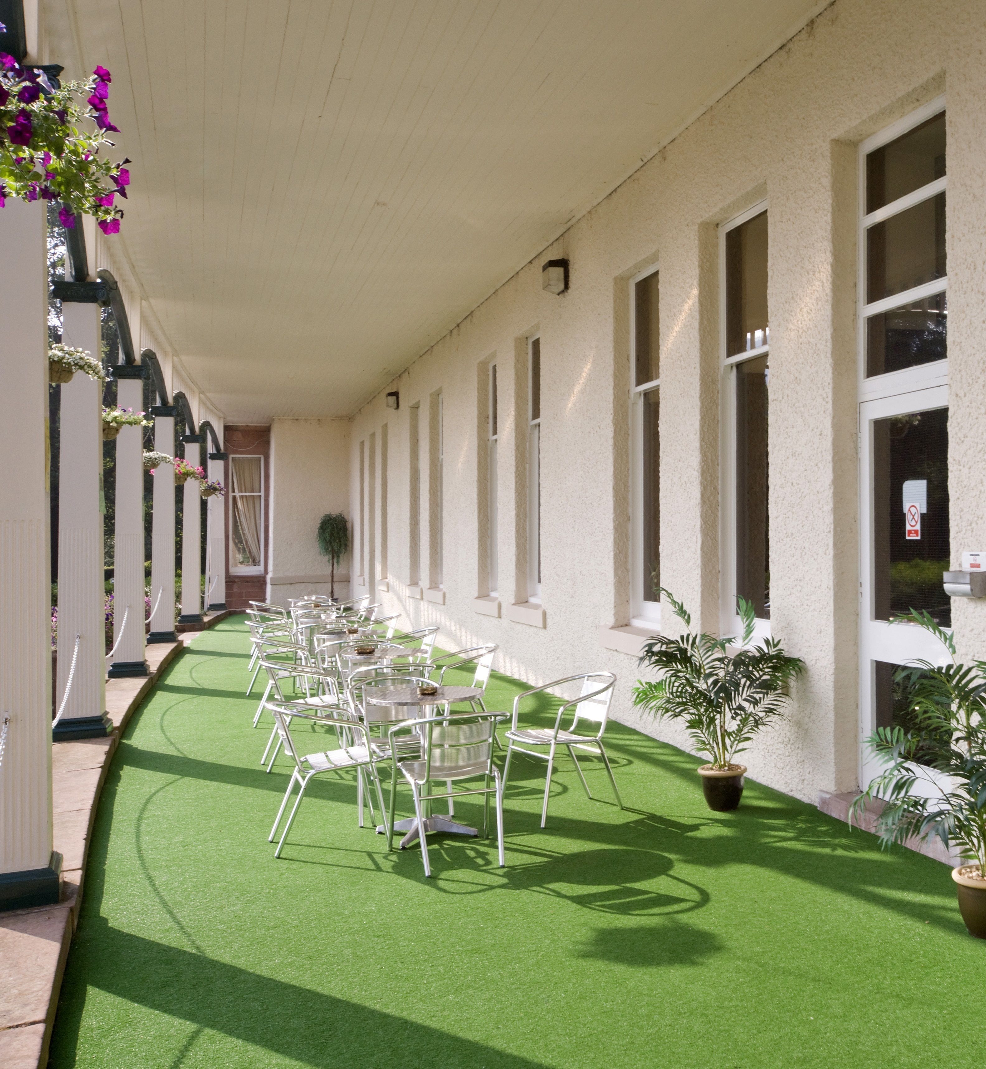 A covered outdoor area with rows of white metal chairs and tables, green artificial turf, and potted plants.