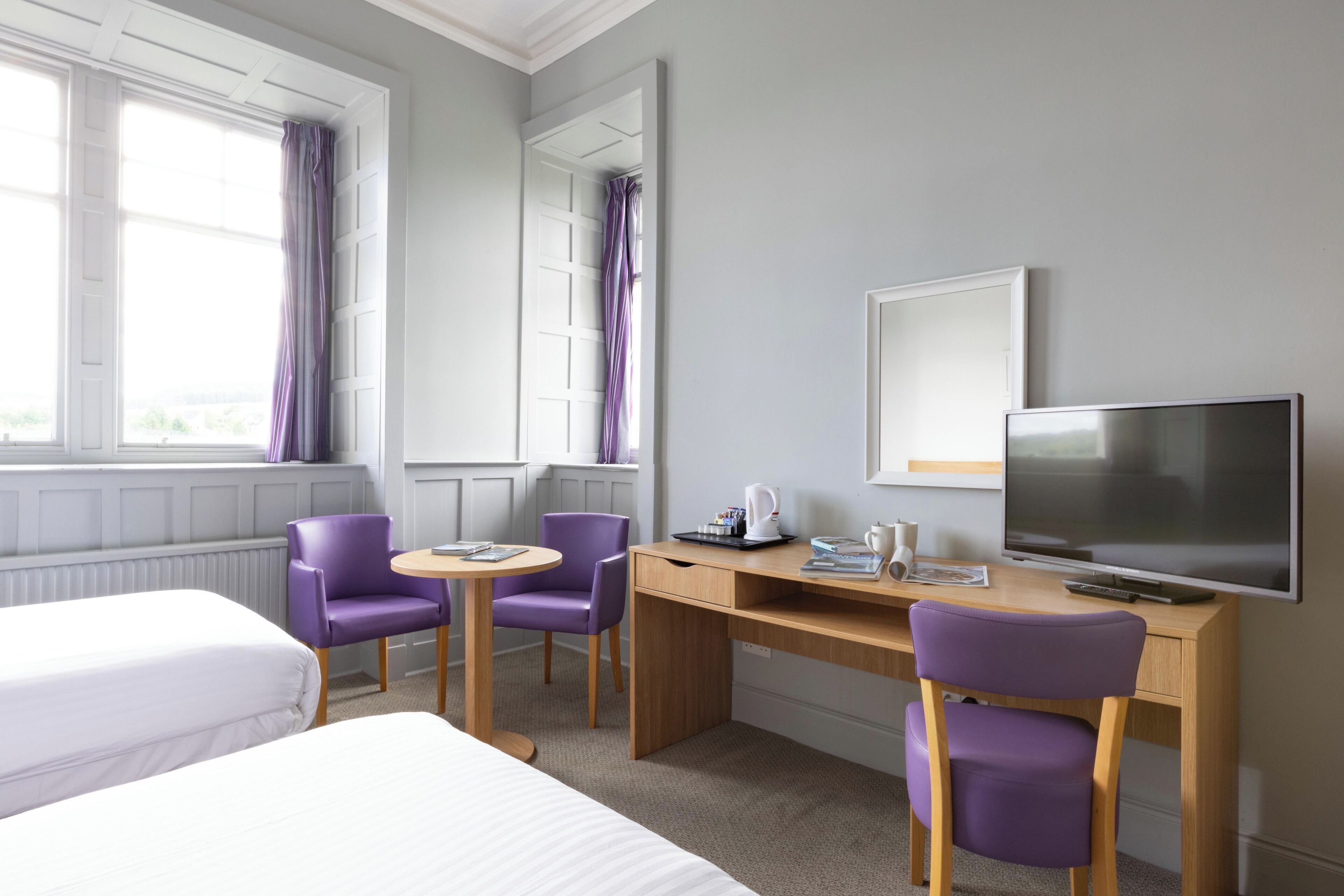 Une chambre d’hôtel avec deux lits, un bureau avec une télévision, une chaise et une fenêtre avec des rideaux.