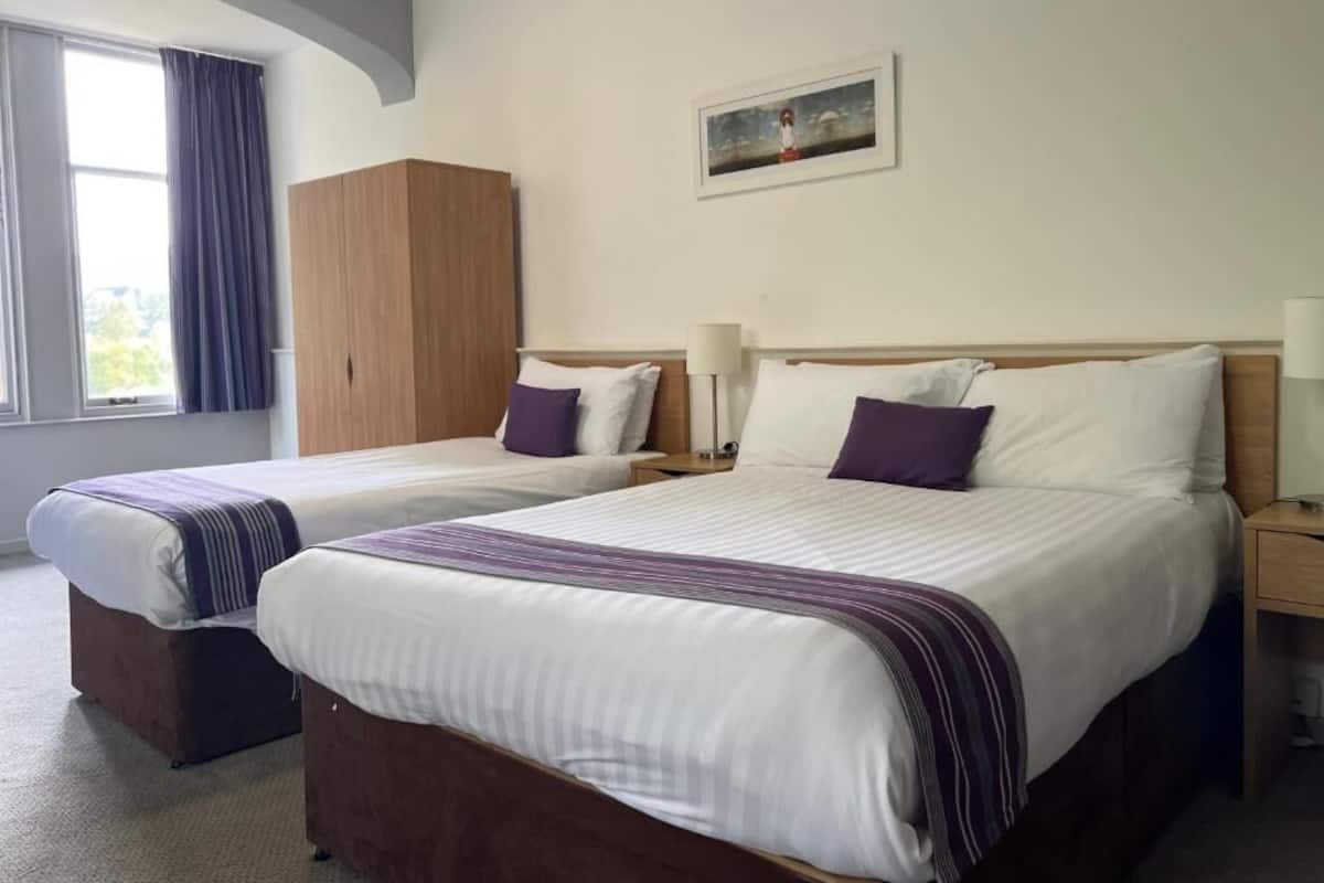 Quarto de hotel com duas camas, guarda-roupa, um quadro na parede e uma janela com cortinas.