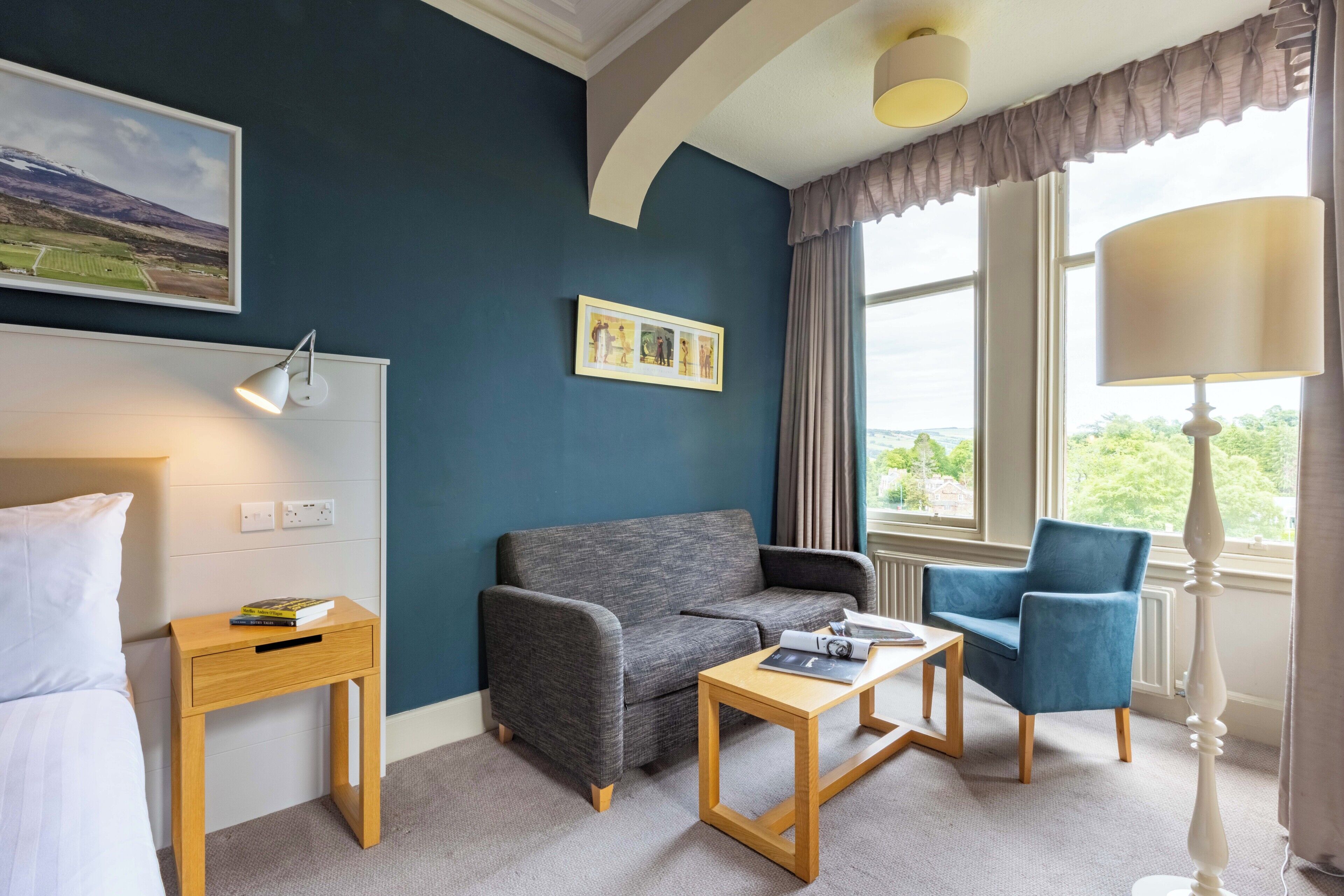 A hotel room with a grey sofa, a blue armchair, a wooden side table, and a lamp.