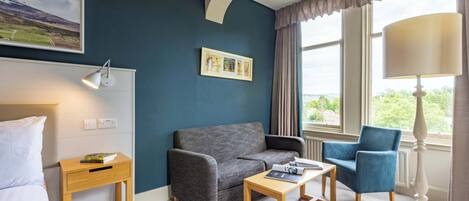 A hotel room with a grey sofa, a blue armchair, a wooden side table, and a lamp.