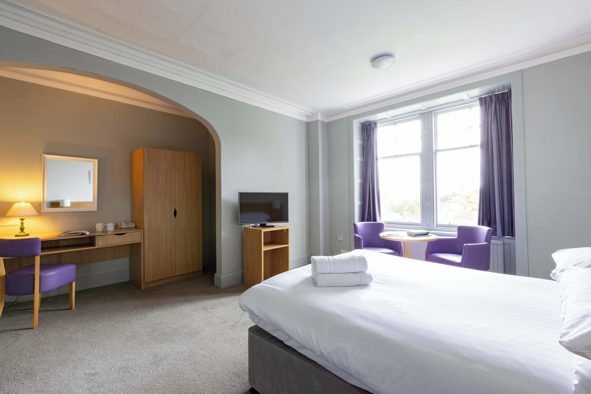 Quarto de hotel com cama, escrivaninha, cadeira, televisão e janela com cortinas.